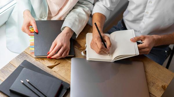 Two people writing in notebooks at a wooden desk with a laptop