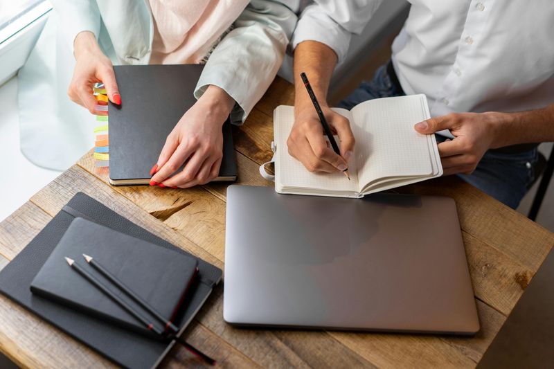 Two people writing in notebooks at a wooden desk with a laptop