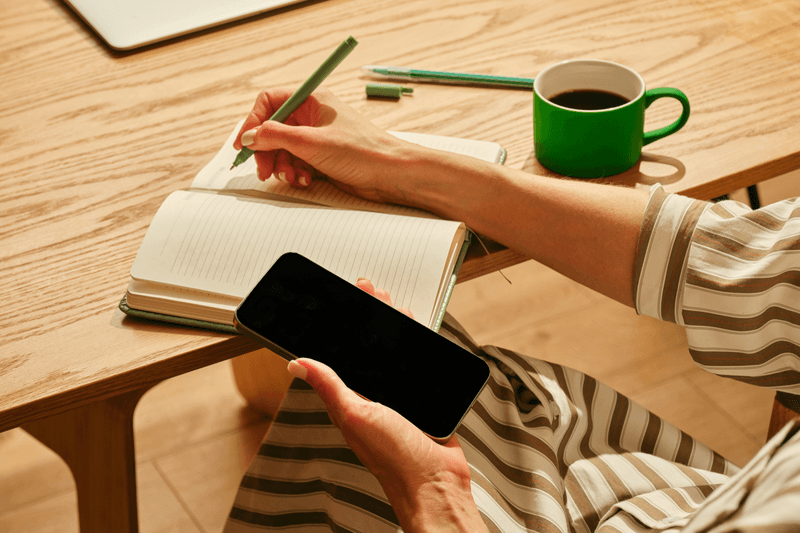 A person writing in a lined notebook with a green pen while holding a smartphone, beside a green mug of coffee on a wooden desk