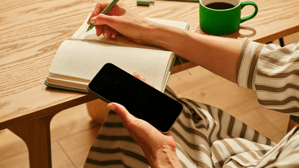 A person writing in a lined notebook with a green pen while holding a smartphone, beside a green mug of coffee on a wooden desk